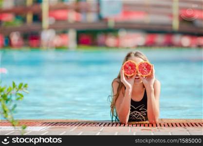 Little girl covers eyes with oranges posing isolated over swimming pool. Little girl covering eyes with orange halves near eyes on background swimming pool
