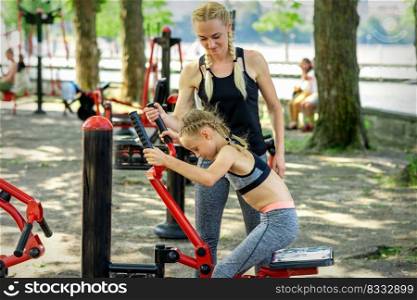 Little girl does exercises on simulator under supervision of young woman coach in the park. Little girl does exercises on simulator