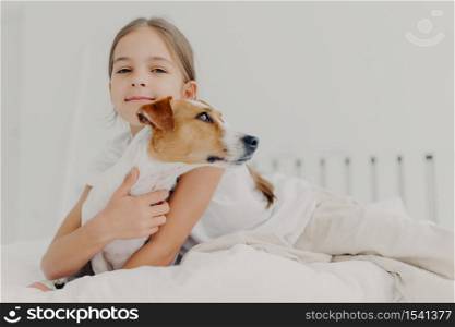 Little girl embraces small pedigree dog, stay in bed, plays with favourite pet before sleep, has appealing look, gazes camly into camera. Childhood and bed time concept. Kid hugs domestic animal