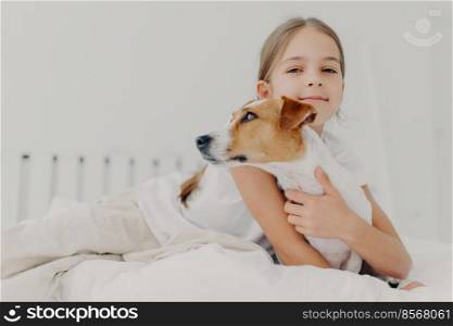 Little girl embraces small pedigree dog, stay in bed, plays with favourite pet before sleep, has appealing look, gazes camly into camera. Childhood and bed time concept. Kid hugs domestic animal