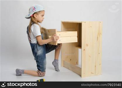 Little girl - furniture collector tries to insert a box in a wooden chest of drawers. Girl in overalls furniture assembler inserts a drawer chest of drawers