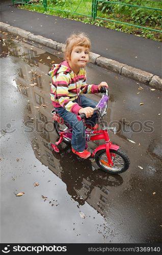 little girl goes on a bicycle on wet asphalt, reflexion in puddle