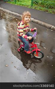 little girl goes on a bicycle on wet asphalt, reflexion in puddle