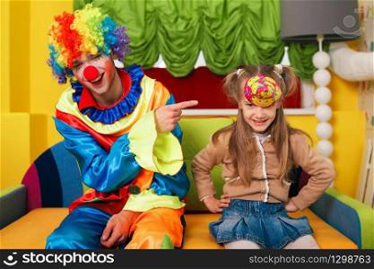 Little girl has put on a cap, and the clown laughs at her. Kindergarten on the background.