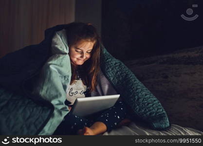 Little girl having fun watching, playing and listening to stories on tablet computer. Child having fun before bedtime laying under duvet using tablet
