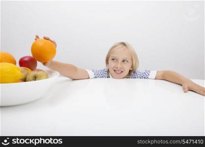 Little girl holding orange from fruit bowl at table