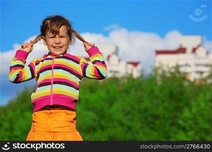 little girl holding plaits in front of trees and buildings