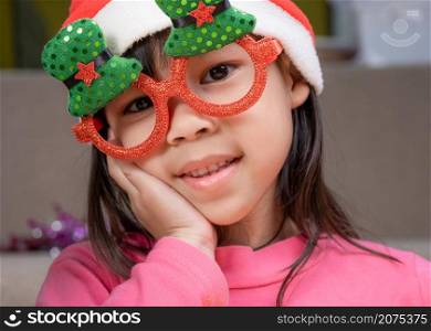 Little girl in a Santa hat sitting on the floor and making wishes near the Christmas tree at home. Happy New Year and Merry Christmas.