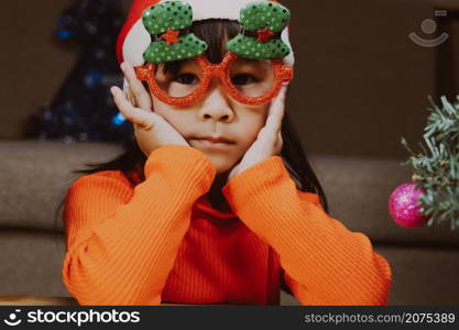 Little girl in a Santa hat sitting on the floor and making wishes near the Christmas tree at home. Happy New Year and Merry Christmas.