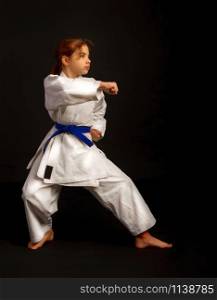 Little girl in a sports kimono and a blue belt performs exercises in kata on a dark background. kata karate girl