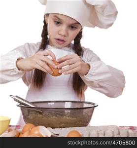 Little girl in a white apron breaks eggs in a deep dish, sharpness on hand and egg