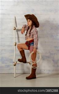 Little girl in a wide-brimmed cowboy hat and traditional dress in high boots and with a lasso posing on a light wooden background