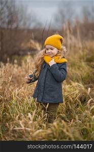 Little girl in a yellow hat and scarf walks along the shore of a river in autumn.. Autumn walk girl in the thickets of red grass 9783.