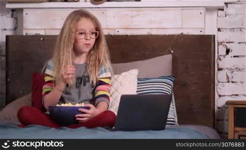 Little girl in glasses with amazing curly blonde hair sitting on the bed in modern style bedroom and eating popcorn while watching cartoons on laptop. Beautiful teen girl watching animated cartoons on modern wireless computer at home.