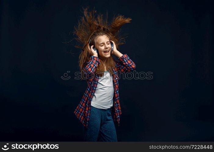 Little girl in headphones against powerful airflow in studio, developing hair effect. Children and wind, kid isolated on dark background, child emotion. Little girl in headphones, developing hair