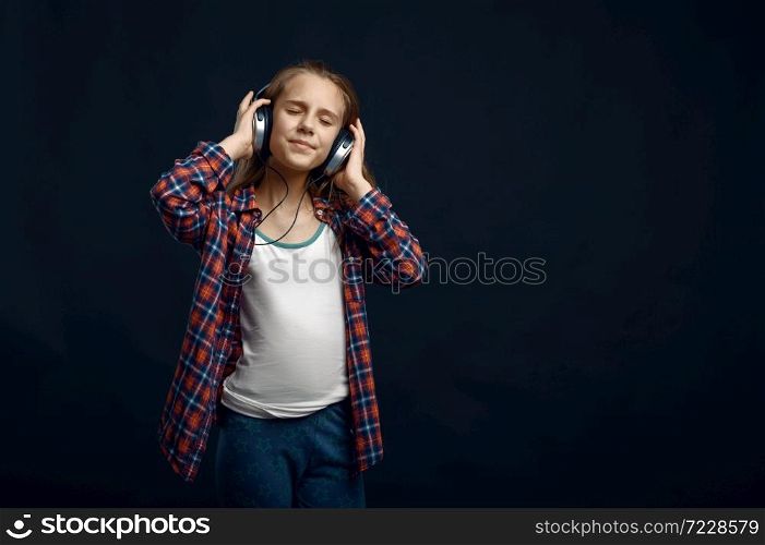 Little girl in headphones against powerful airflow in studio, developing hair effect. Children and wind, kid isolated on dark background, child emotion. Little girl in headphones, developing hair
