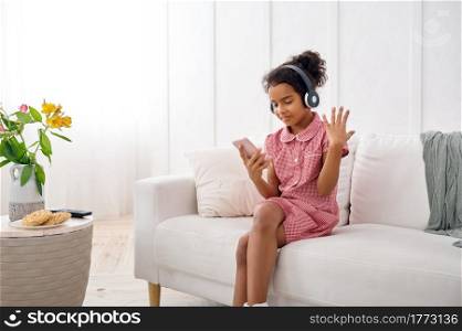 Little girl in headphones listening to music at home. Female kid sitting on sofa and using mobile phone, funny child with modern gadget. Little girl in headphones listening to music