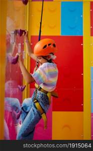 Little girl in helmet poses on climbing wall, entertainment center, young climber. Children having fun, kids spend the weekend on playground, active childhood. Little girl in helmet poses on climbing wall