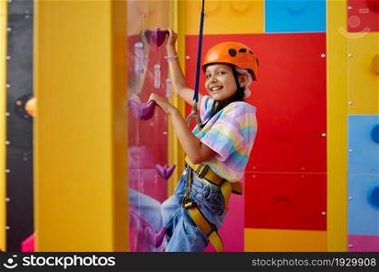 Little girl in helmet poses on climbing wall, entertainment center, young climber. Children having fun, kids spend the weekend on playground, happy childhood. Little girl in helmet poses on climbing wall