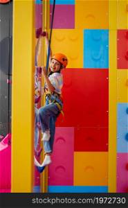 Little girl in helmet poses on climbing wall, entertainment center, young climber. Children having fun, kids spend the weekend on playground, active childhood. Little girl in helmet poses on climbing wall