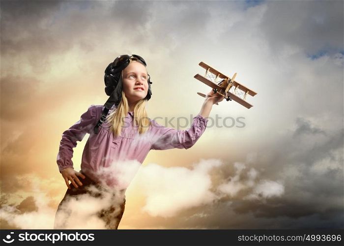 Little girl in pilot&rsquo;s hat. Image of little girl in pilots helmet playing with toy airplane against clouds background
