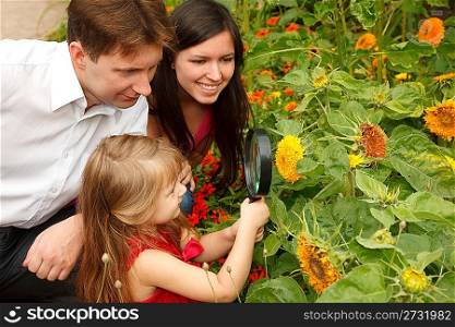 Little girl in red dress considers flower together with parents through magnifying glass.