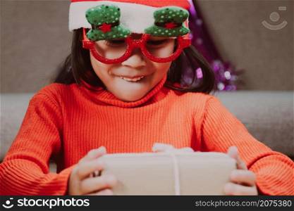 Little girl in Santa hat opening gift box sitting in the living room with Christmas tree at home. Happy New Year and Merry Christmas.