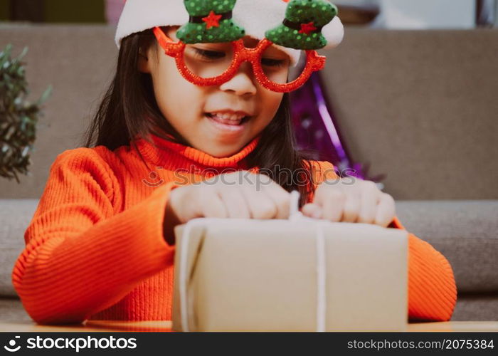 Little girl in Santa hat opening gift box sitting in the living room with Christmas tree at home. Happy New Year and Merry Christmas.