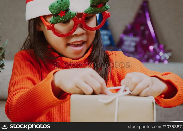 Little girl in Santa hat opening gift box sitting in the living room with Christmas tree at home. Happy New Year and Merry Christmas.