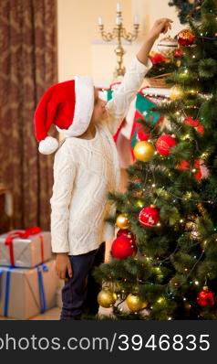 Little girl in Santa&rsquo;s hat putting golden ball on top of Christmas tree