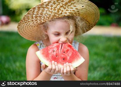 Little girl in straw hat with big slice of watermelon sitting on green grass in summer park.. Little girl in straw hat with big slice of watermelon sitting on green grass in summer park