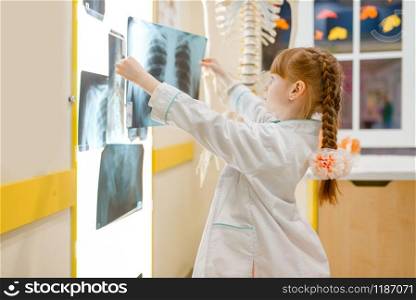Little girl in uniform looks at the x-ray, playing doctor, playroom. Kid plays medicine worker in imaginary hospital, profession learning. Little girl in uniform looks at the x-ray, doctor