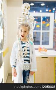 Little girl in uniform near a human skeleton, playing doctor, playroom. Kid plays medicine worker in imaginary hospital, profession learning, childish dream