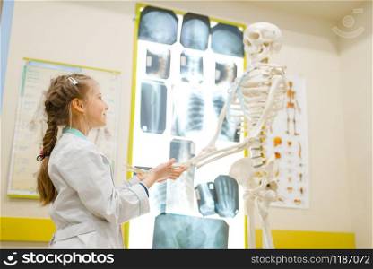 Little girl in uniform playing doctor with human skeleton, playroom. Kid plays medicine worker in imaginary hospital, profession learning, childish dream