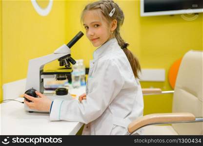 Little girl in uniform sitting at the microscope and playing doctor, playroom. Kid plays medicine worker in imaginary hospital lab, profession learning, childish dream