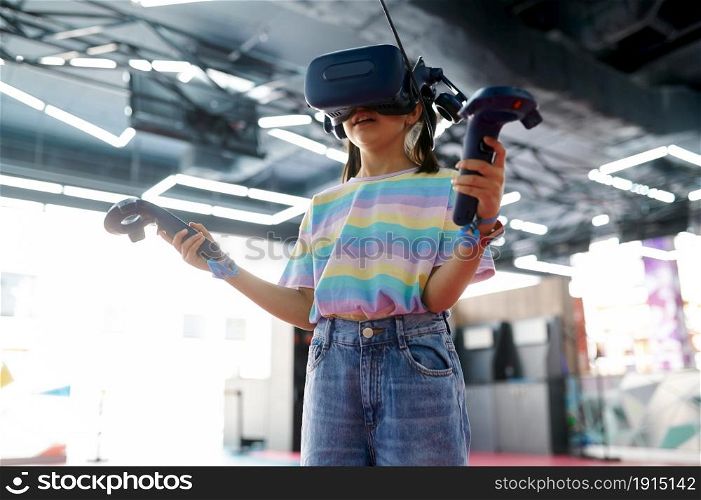 Little girl in virtual reality glasses holds joysticks in her hands. Kid playing 3d video game in entertainment center. Children having fun, child spend the weekend on playground, happy childhood. Little girl in virtual reality glasses, playground