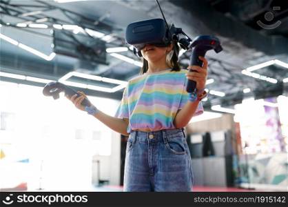 Little girl in virtual reality glasses holds joysticks in her hands. Kid playing 3d video game in entertainment center. Children having fun, child spend the weekend on playground, happy childhood. Little girl in virtual reality glasses, playground