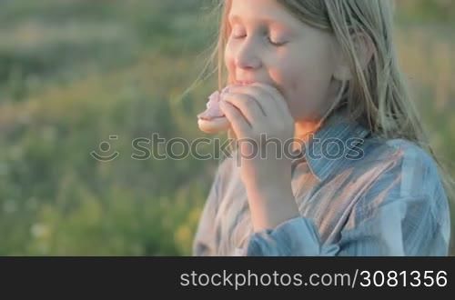 Little girl is eating a sandwich, smiling and looking at the sunset. Close-up.