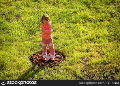 little girl is standing and frowning on water drain hatch in grass field.