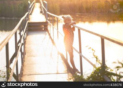 Little girl is standing on the bridge over the river and looking at the camera in sunset light