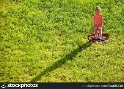 little girl is standing on water drain hatch in grass field. girl in right upper corner. long shadow from girl