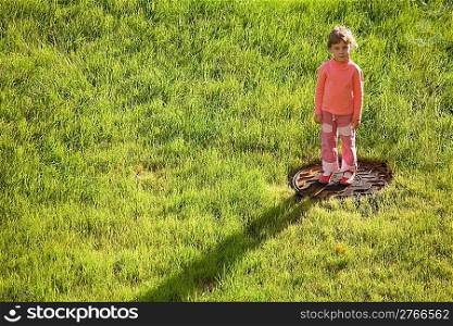 little girl is standing on water drain hatch in grass field. long shadow on grass
