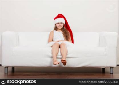 Little girl kid in santa claus hat. Christmas.. Cute little girl kid in red santa claus hat and white dress sitting on sofa couch. Chrtistmas holiday season.
