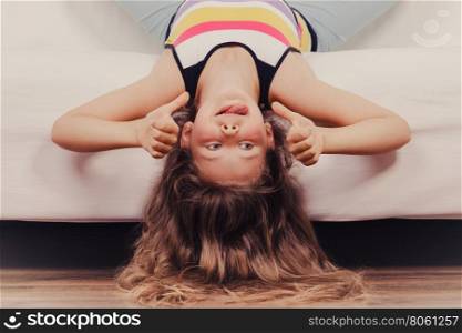 Little girl kid with long hair upside down on sofa. Little girl with long hair lying upside down on sofa at home. Kid playing having fun on couch giving thumb up gesture.