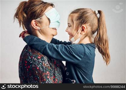 Little girl kissing her mother who has covered eyes with face mask for fun. Daughter giving kiss her mom during covid-19