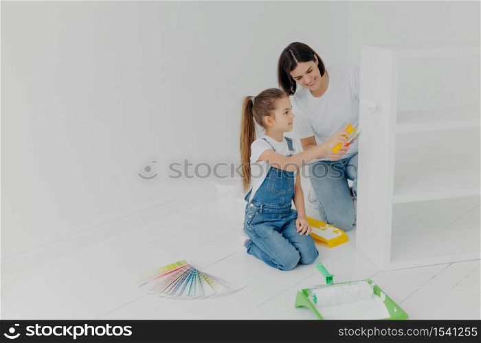 Little girl learns how to paint with roller, stands on knees, her mother tells how to do it correctly, pose in spacious white room, choose color from palette samples. Home makeover, decoration concept