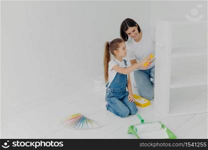 Little girl learns how to paint with roller, stands on knees, her mother tells how to do it correctly, pose in spacious white room, choose color from palette samples. Home makeover, decoration concept