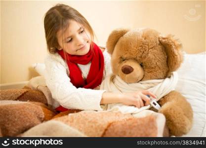 Little girl lying in bed and measuring teddy bears temperature with thermometer