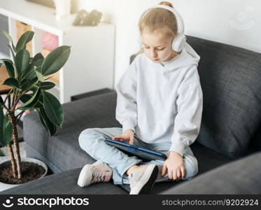 Little girl  lying on the couch with an electronic tablet listens to music in white headphones. Using wireless devices for education.