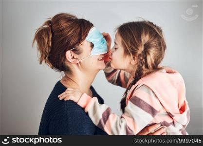 Little girl moving her mom’s face mask on eyes and giving a kiss her mother. Funny moments during quarantine and covid-19 pandemic